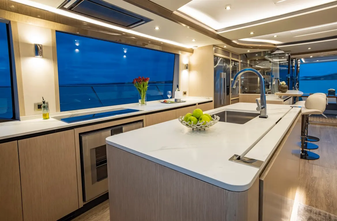 Modern yacht kitchen with marble countertops, a bowl of green apples, and large windows showcasing a twilight sea view.